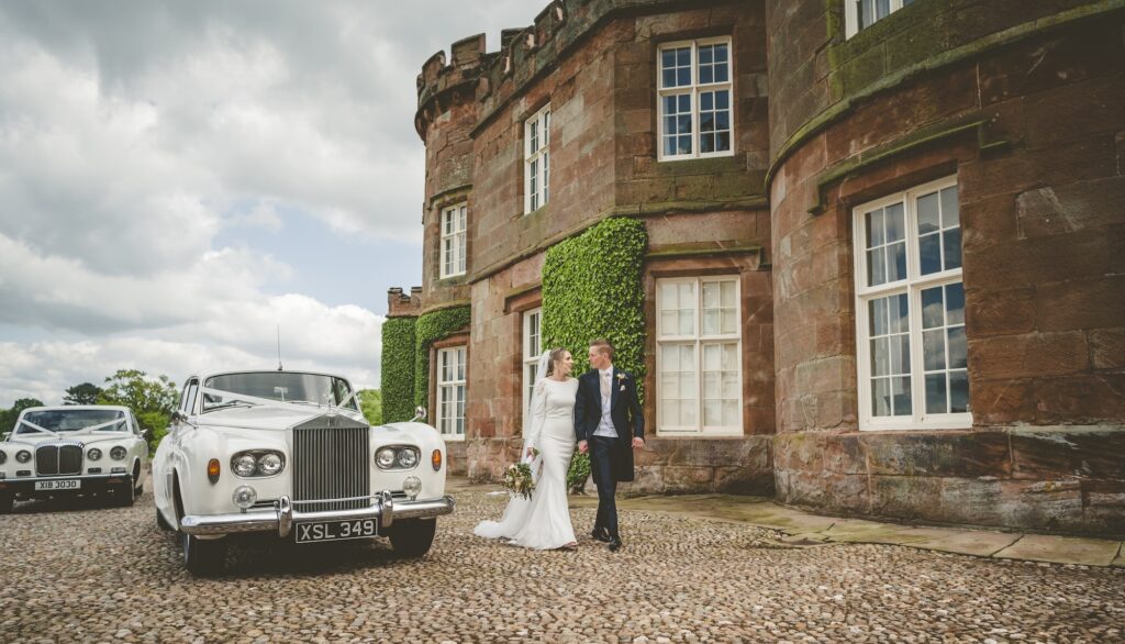 Bride and groom walking beside a classic Rolls-Royce outside The Citadel's castle-style country house before their Shropshire marquee wedding